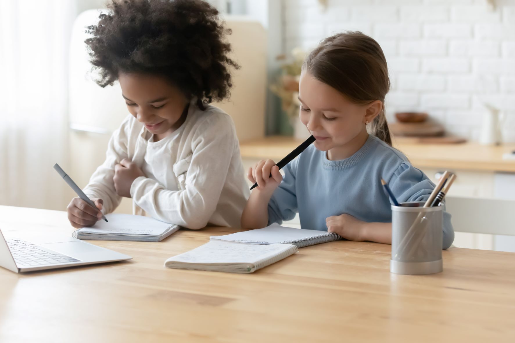 Two smiling little girls sitting next to each other at a table, writing in notebooks.