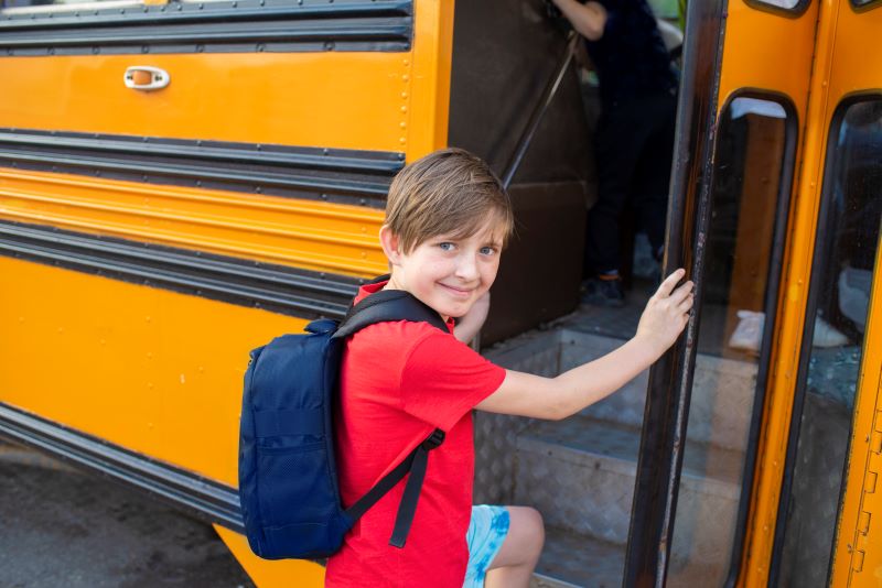 A smiling middle school-aged boy with a blue backpack getting on a yellow school bus.