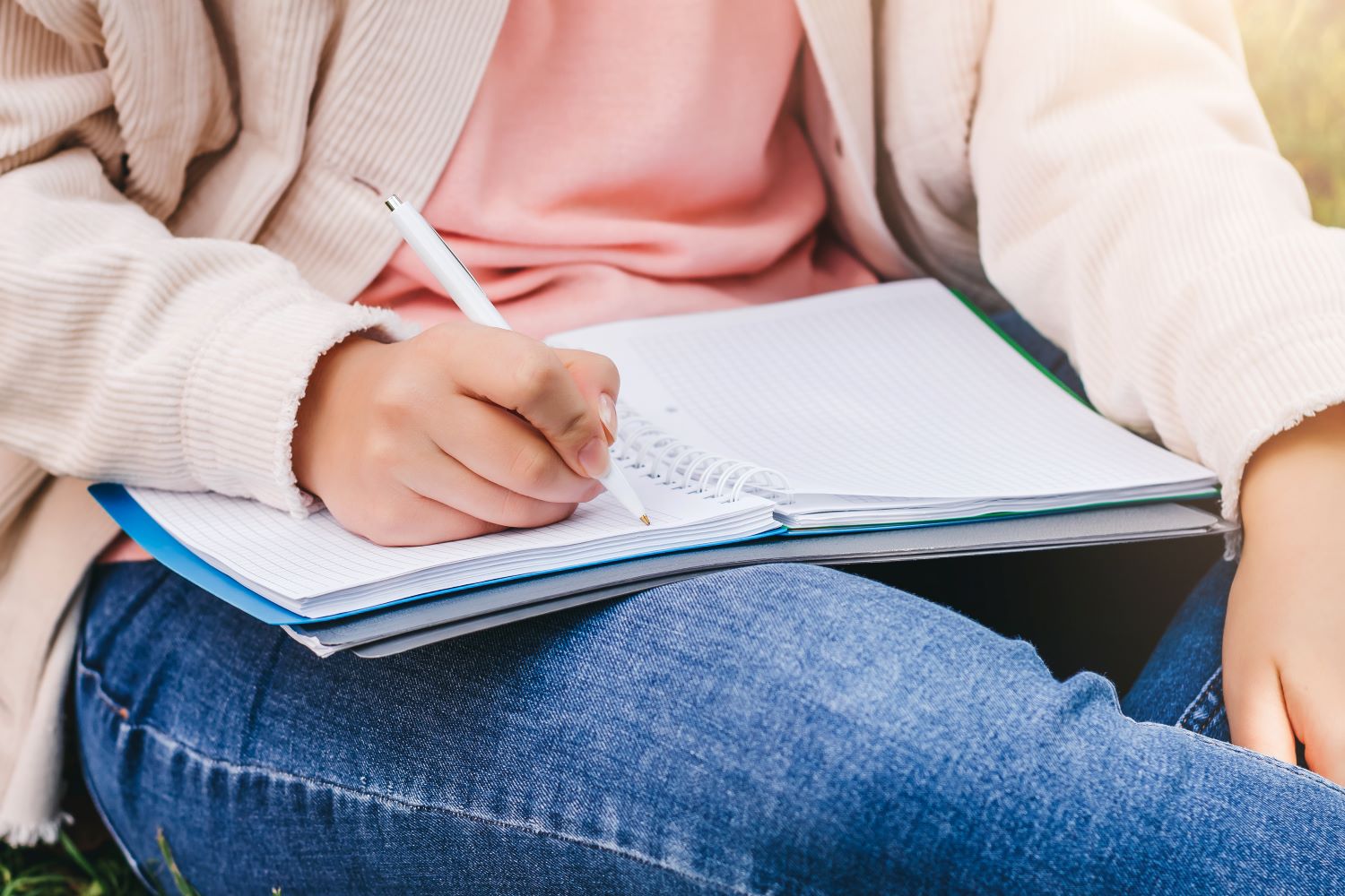 A close-up of a girl sitting in the grass writing in a journal.