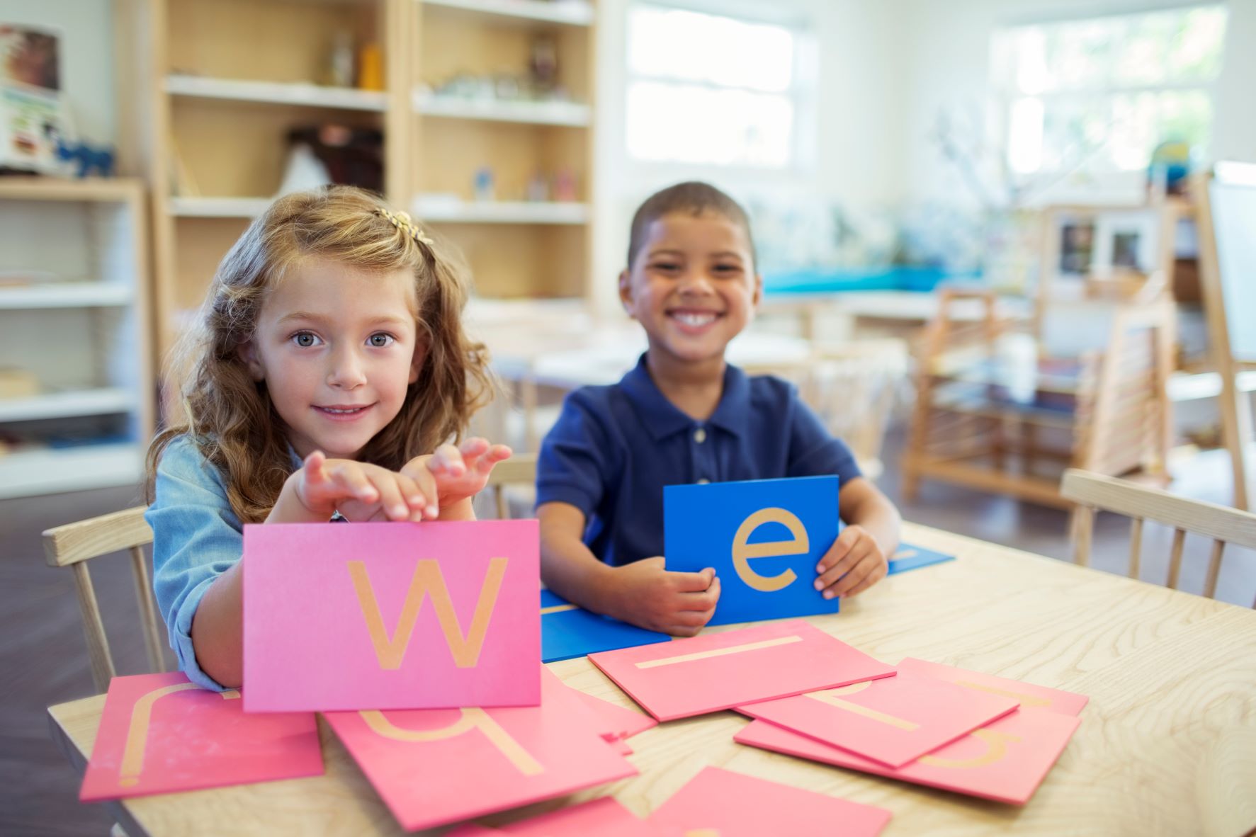 A little girl and little boy, smiling, in a classroom holding up the letters W and E.