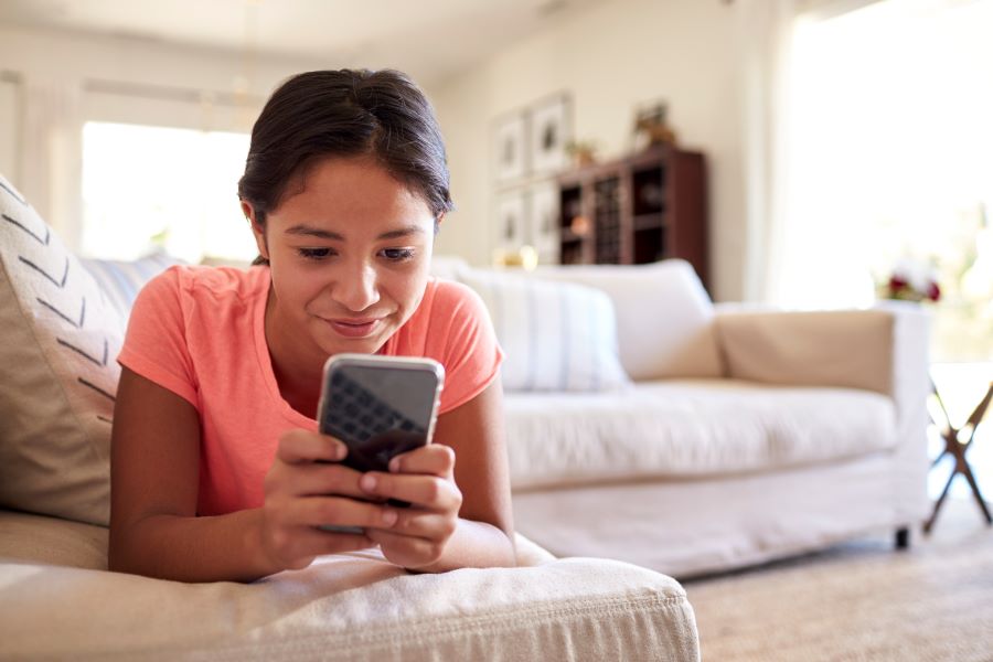 A middle school-age girl lying on her stomach on the couch smiling at her phone in her hands.