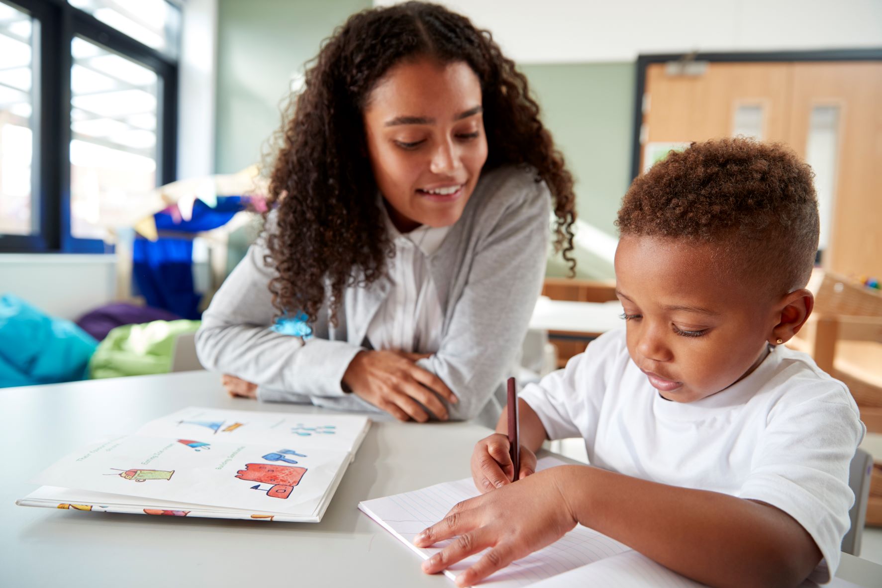 A teacher sits with a young student while he writes on a piece of paper.