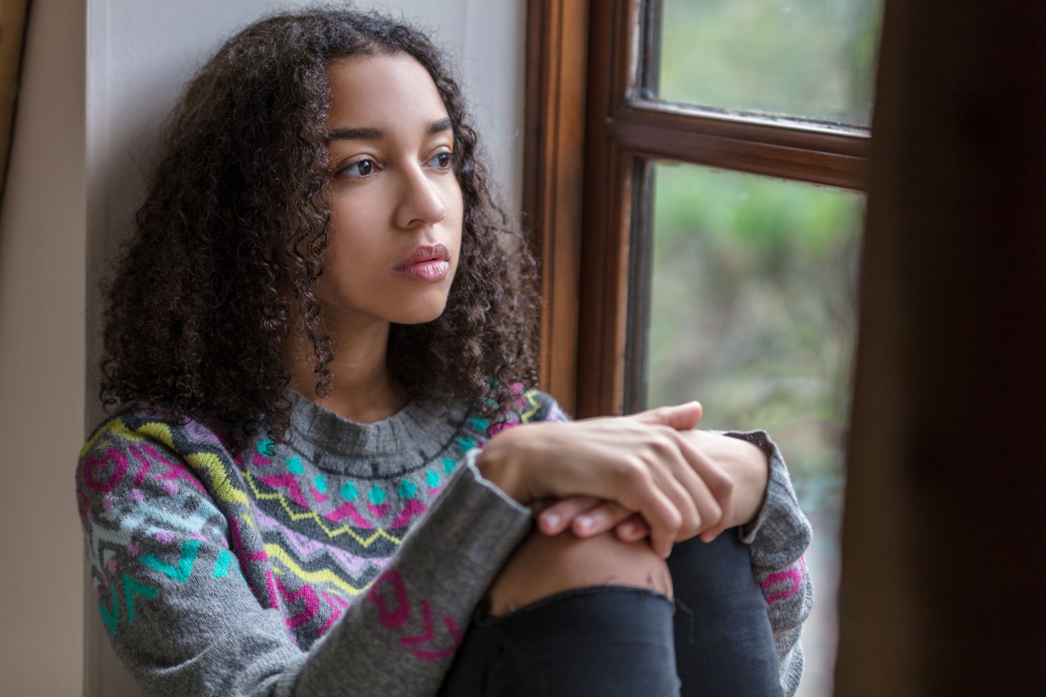 A sad-looking teen girl sits looking wistfully out a window.