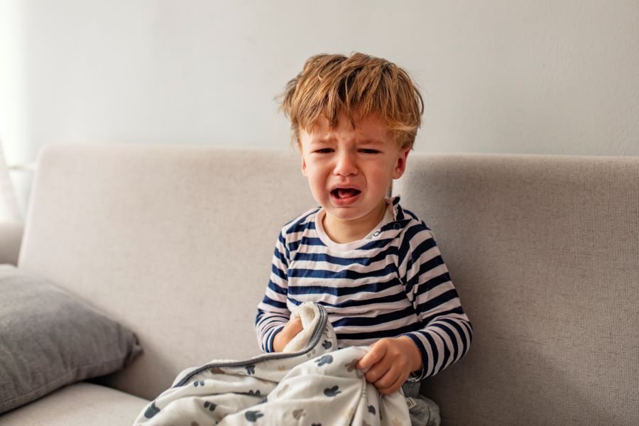 A little boy sitting on a couch with a blanket on his lap, crying.