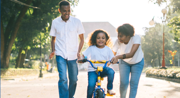 homepage-video-placeholder Girl on bike with parents helping.