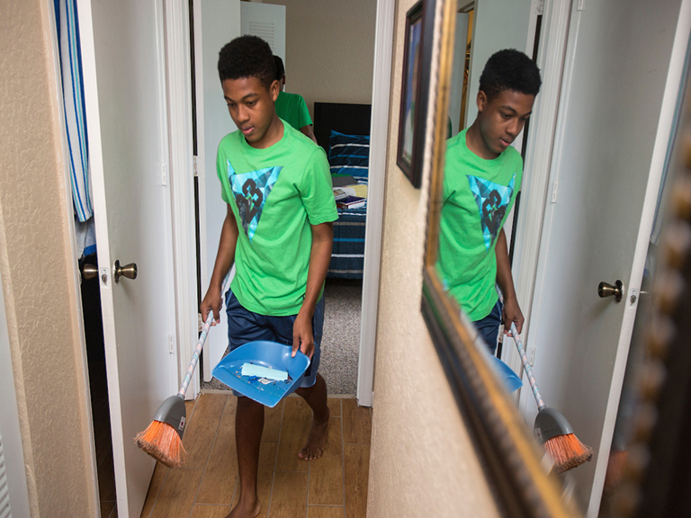 Teen boy walks down a hallway in his home with a broom and dustpan.