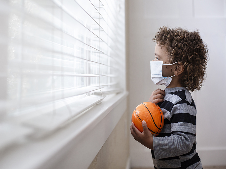 Little curly headed boy with mask on holding a small basketball looking anxiously out the window from inside a home.