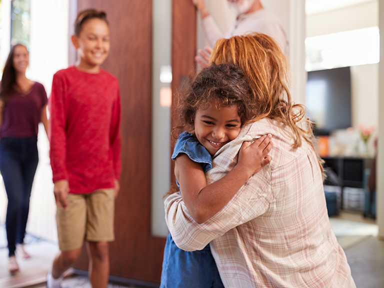 Granddaughter smiling and hugging grandmother, while mother and grandson walk through the door to greet grandmother.