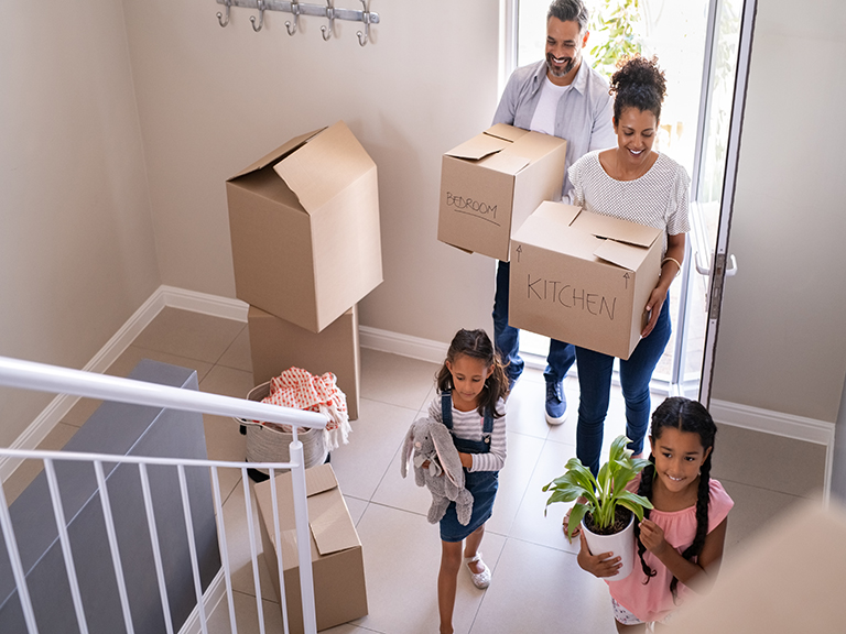 Mother and family holding boxes enter their new home, with two little girls running excitedly ahead.