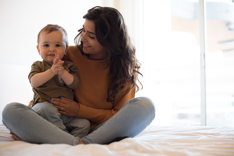 Mother sitting cross-legged on the floor playing with her baby, who's smiling and clapping while sitting on her lap.