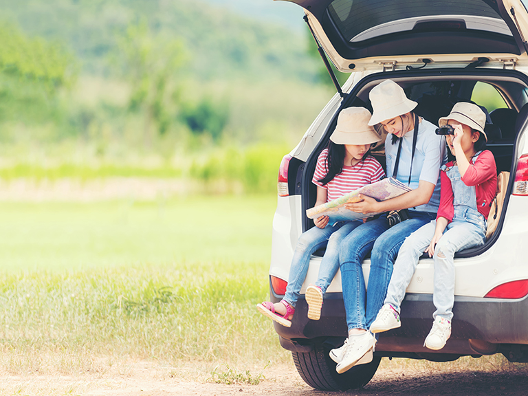 Mom and two daughters sit in an open trunk of their car, looking at a map. One girl has binoculars and is looking at the view.