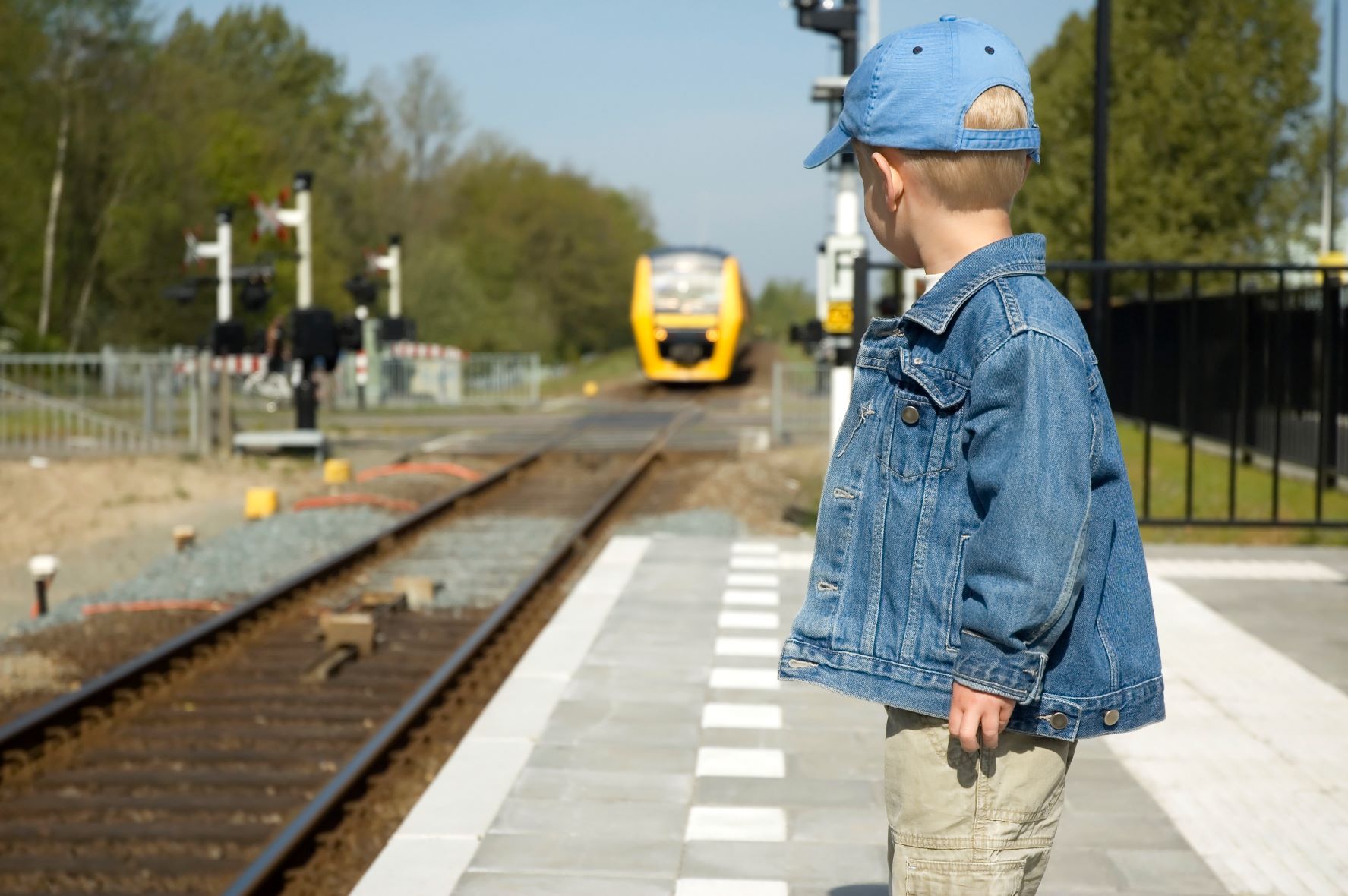 A little boy in a denim hat and jacket standing at a cross walk watching a yellow training coming down the tracks.