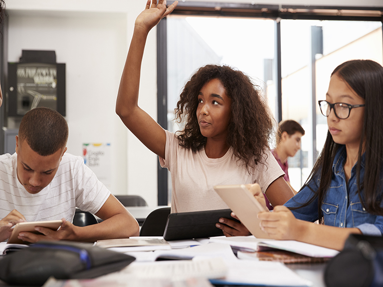 Teenage girl raises her hand while sitting at a table in class, surrounded by other students.
