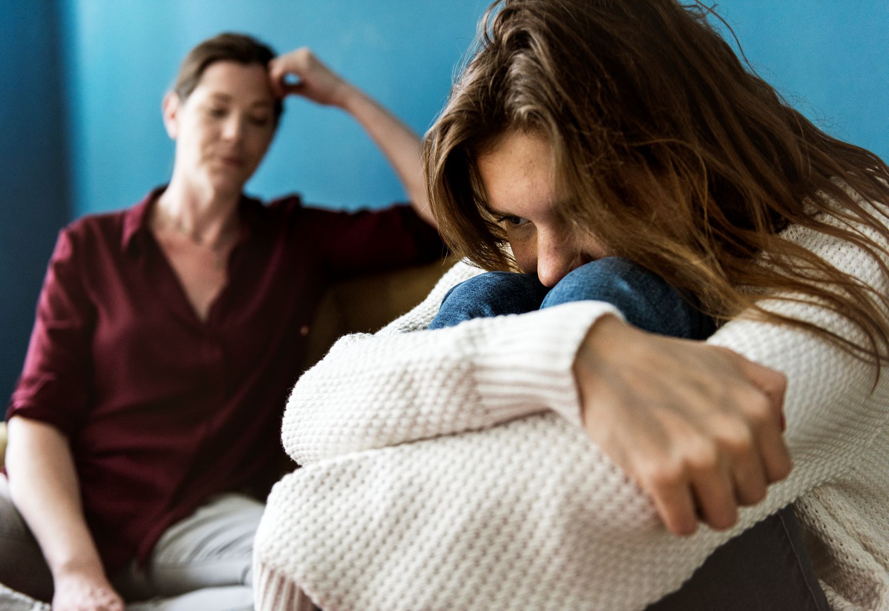 Teen girl with long hair covering face curled up on couch, face buried in her knees, after a fight with her mom, who's in the background.