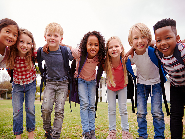 Young children of different races outside in a circle with their arms around each other.