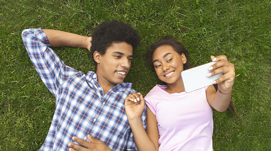 Teenage boy and girl laying in the grass, smiling and taking a selfie with a cellphone.