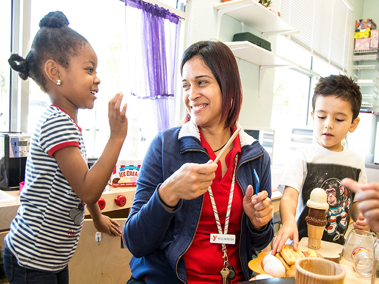 A preschool teacher smiles at a little girl while doing arts and crafts with the girl and other children at a table in a classroom.
